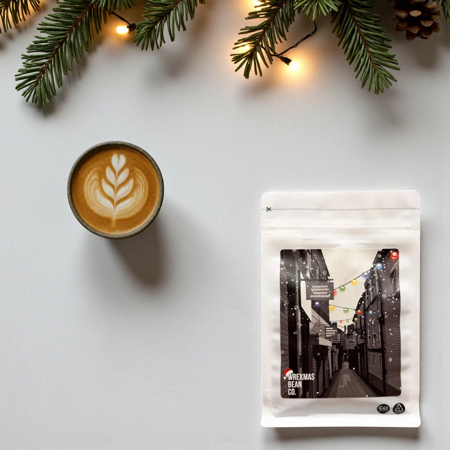 Top-down view of the Wrexham Bean Christmas Blend coffee bag beside a cup of black coffee with soft festive greenery and warm lights.